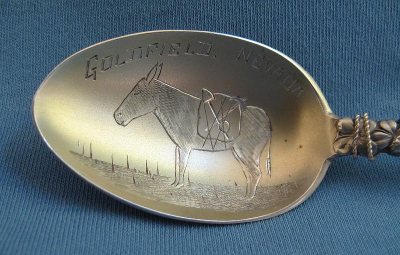 Souvenir Mining Spoon Bowl Goldfield, NV.JPG - SOUVENIR MINING SPOON GOLDFIELD NV - Sterling silver spoon with engraved scene of miner’s pack mule in gold washed bowl, marked GOLDFIELD,NEVADA, ca. 1905, back marked Sterling with maker’s mark, made by Joseph Mayer & Bros., Seattle, WA 1895-1945, figural handle with wire wrap has miner’s gold pan with nuggets and pick and shovel marked NEVADA in pan on handle and double-sided pack mule on top, length 5 5/8 in. and weight 24 g  [Goldfield is an unincorporated community and the county seat of Esmeralda County, Nevada. It had a resident population of 268 in the 2010 census and is located 247 miles southeast of Carson City.  Goldfield was a boomtown in the first decade of the 20th century due to the discovery of gold.  The town was the site of a rare post-1900 major gold discovery, unusual in that most major discoveries in the lower 48 occurred before 1880. The ore initially was very rich, spurring rapid growth of the town from 1902 until 1906. The Goldfield Mining District was discovered in late 1902 and the Combination Lode was located in 1903 and the major rush followed.  From 1903 to 1910, Goldfield was the largest city in Nevada, peaking at 30,000 residents in 1908, the year the Goldfield Consolidated Mines Company erected a 100-stamp mill. The town was called the "Queen of the Mining Camps" for its luxury and availability of saloons and other forms of entertainment, including its many sporting houses. During this time, building lots often sold for as much as $45,000. In addition to its numerous saloons, the town boasted three newspapers, five banks and a mining stock exchange. Like so many boomtowns, labor difficulties soon followed.  After mining on an extensive scale began, the miners organized themselves as a local branch of the Western Federation of Miners. The Goldfield Consolidated Mines Company, primarily owned by George Wingfield, held a virtual monopoly in the town which led to an adversarial relationship between mine owners and the unions.  There were several strikes in December 1906 and January 1907 for higher wages, with more strikes continuing throughout the year for various reasons.  Ultimately federal troops were stationed in Goldfield from December 1907 to March 1908.  Goldfield experienced one of the most dramatic rises and subsequent crashes of all the mining towns of the Old West.  By 1908 ore production was already in steep decline and by 1910, the population of Goldfield dropped below 5,000.  In 1912, gold production had dropped from its former high of 11 million dollars per year to 5 million dollars and by 1918, the mines produced only 1 ½ million dollars in ore, with half that amount in the next year. By 1920, the town was called home to only about 1,500 residents and for the next three years, only a cumulative $150,000 in ore was produced by the area mines.  Much of the town was destroyed by a fire in 1923, although several buildings survived and remain today, notably the Goldfield Hotel, the Consolidated Mines Building, and the schoolhouse.  Between 1903 and 1940, Goldfield's mines produced more than $86 million.  Gold exploration continues in and around the town today.]